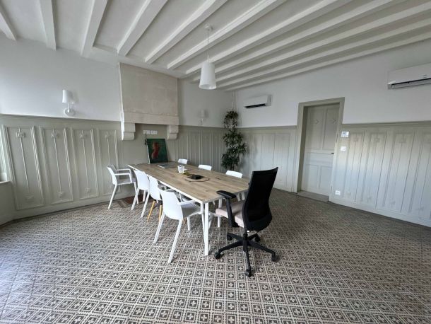 Salle de réunion élégante avec une grande table en bois, entourée de chaises blanches et d’un fauteuil ergonomique noir, dans un espace avec un sol en carreaux de ciment et des murs moulurés, éclairé par des suspensions modernes.