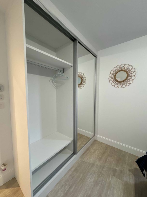 Placard sur mesure avec portes coulissantes, une en miroir et l'autre en finition blanche, équipé d'une penderie et d'une étagère, installé dans une pièce moderne avec un sol en parquet clair.